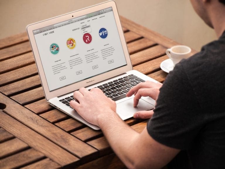 Man using a laptop showing a website interface, seated at a wooden table with a coffee cup beside him.
