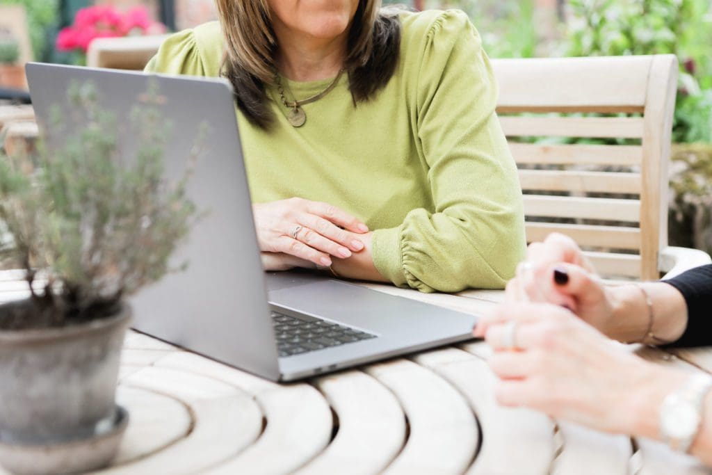 Woman in green top using laptop at garden table during a digital marketing client kickoff meeting with another person partially visible.