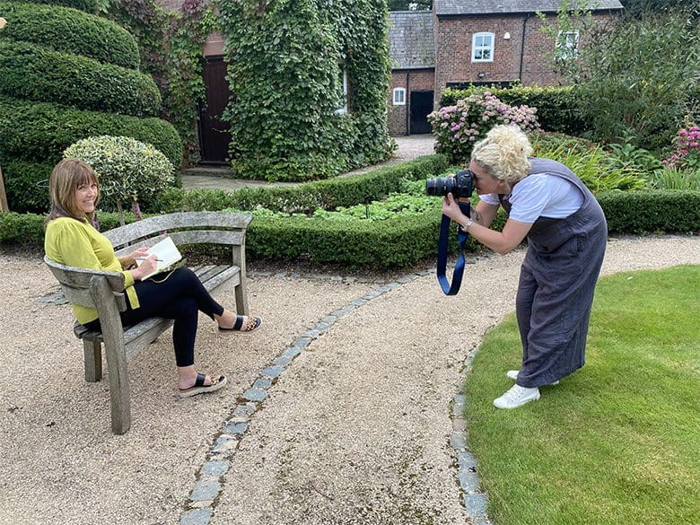 A woman sitting on a garden bench reading a book while another woman takes her photograph for a brand shoot in a lush garden setting.
