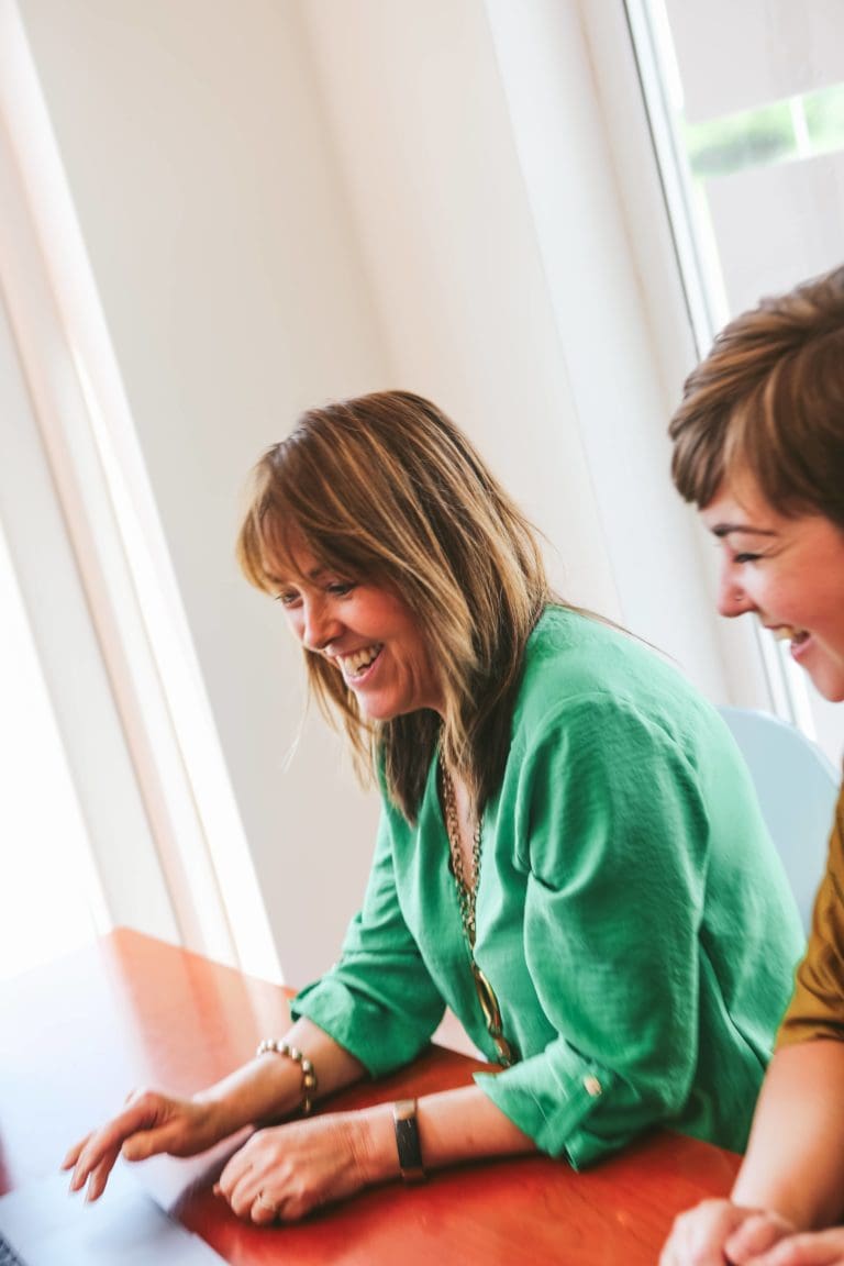 Woman in green top using laptop at meeting table during a digital marketing client kickoff meeting with another woman.