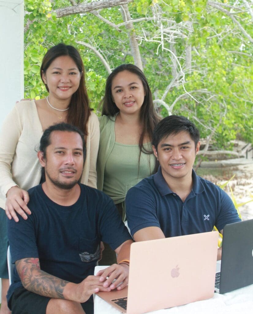 Four people sit outdoors at a table with a laptop, discussing website updates. Two are seated in front and two stand behind them. They are smiling, and greenery can be seen in the background.