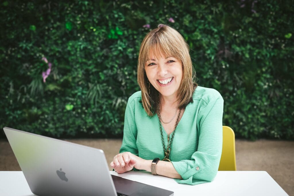 Jeanette in a green shirt sits at a table in front of a laptop, smiling as she answers a contact form submission. There is a vibrant green wall with foliage in the background.