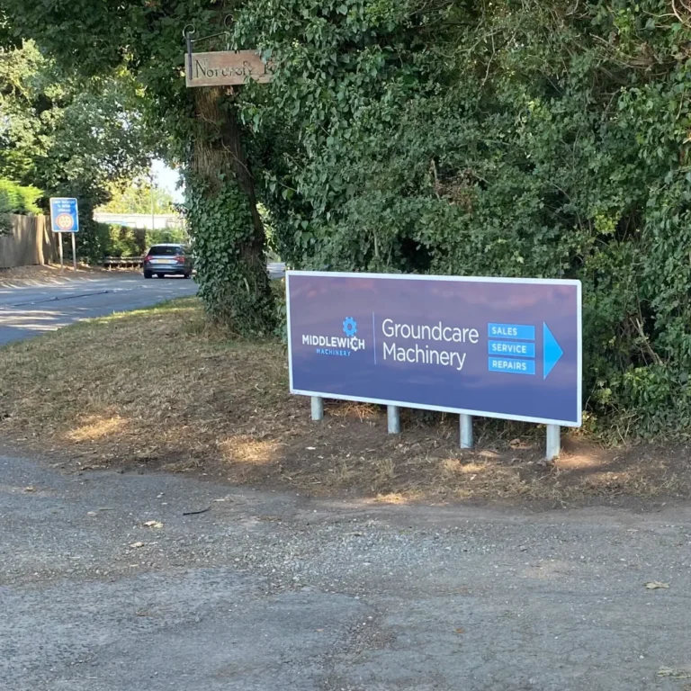 A roadside sign for Middlewich Machinery directing to Groundcare Machinery sales, service, and repairs. Signage clearly visible as a car drives along the tree-lined road.
