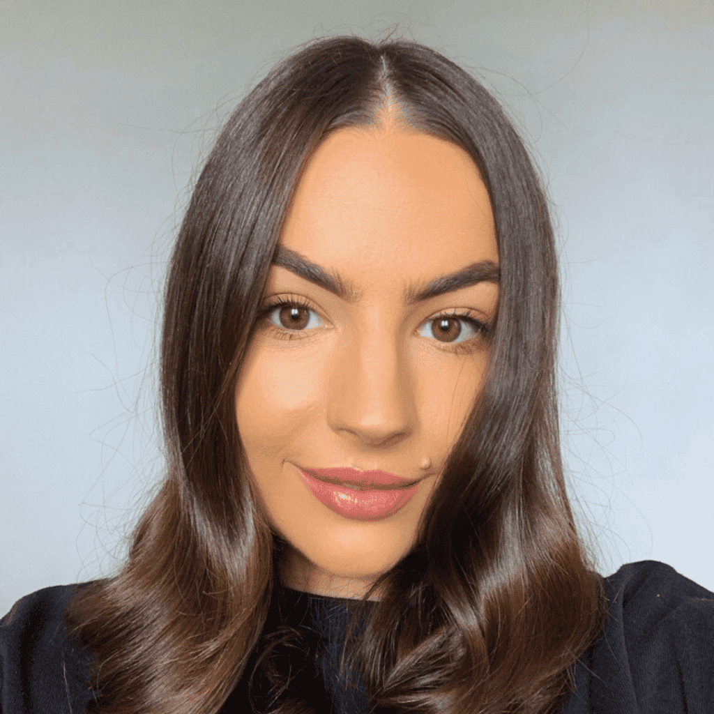 Close-up of a woman with long brown hair, styled in waves. She has arched eyebrows and wears subtle makeup with a hint of pink lip color. The plain, light-colored background accentuates her features beautifully, giving insight into more about her elegant style.