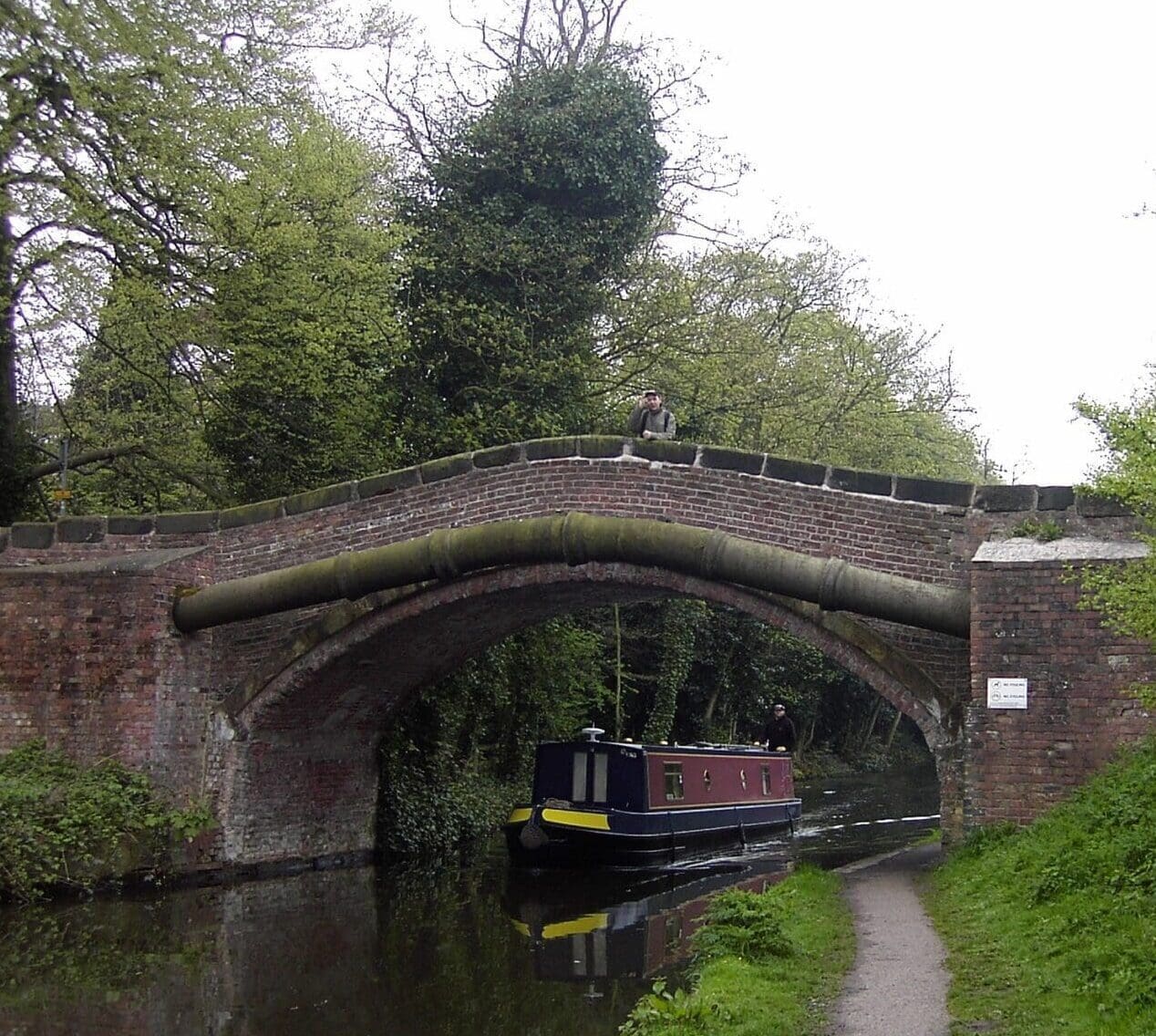 A narrowboat glides under a brick arched canal bridge, flanked by lush greenery. To the right, a footpath meanders alongside, possibly leading to quaint service areas that offer charming rest stops amidst the serene waterscape.