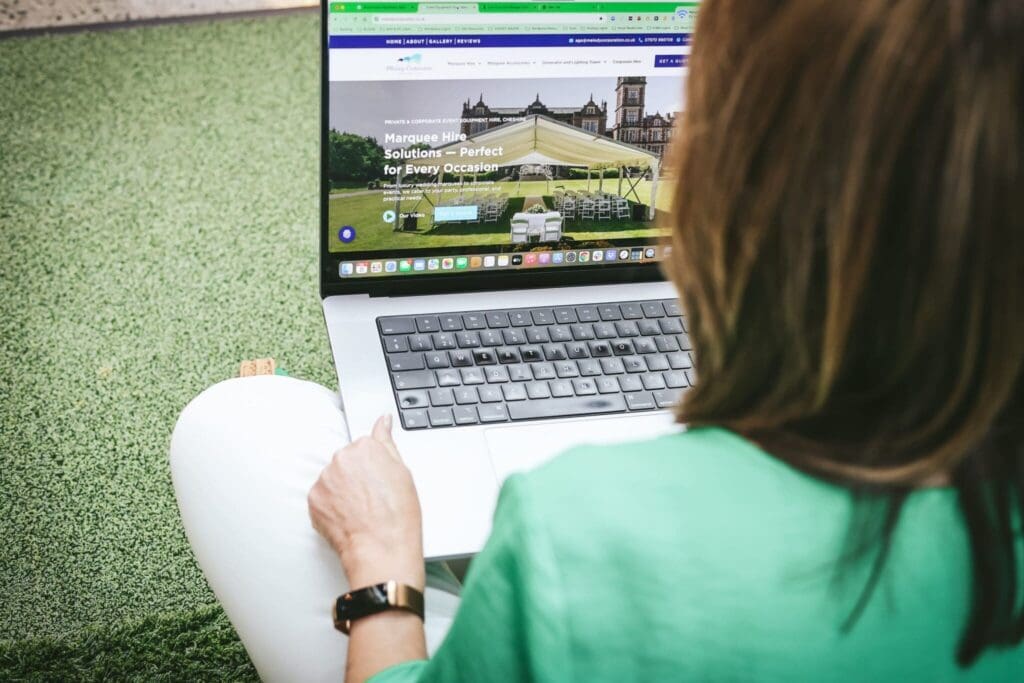 Person in a green shirt using a laptop on a grassy surface, viewing a website about marquee hire services.