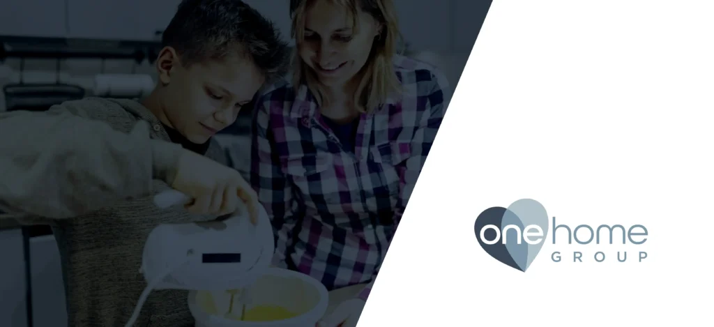 A child uses a hand mixer while an adult watches, both smiling in a kitchen. To the right, the heart-shaped One Home Group logo is displayed on a white background.
